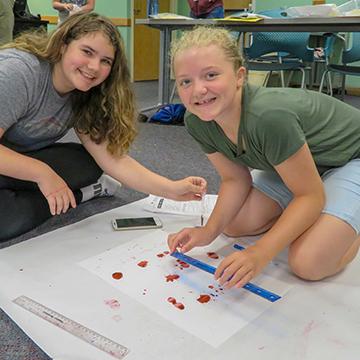 two female students sit on the floor while measuring on a large piece of white paper